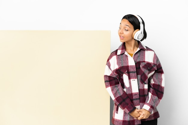 Mujer joven escuchando música con un gran cartel vacío sobre fondo aislado. Retrato