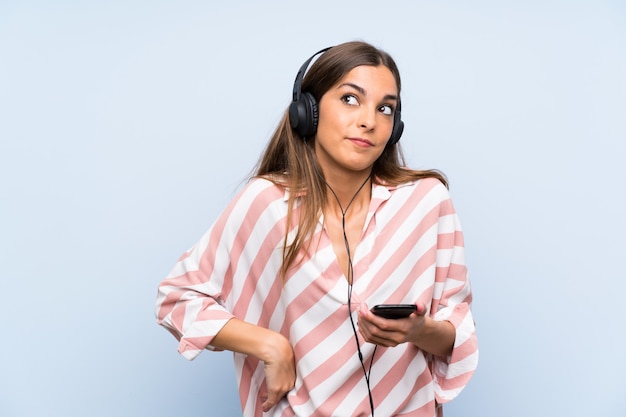 Mujer joven escuchando música con un móvil sobre pared azul aislada haciendo dudas gesto mientras levanta los hombros