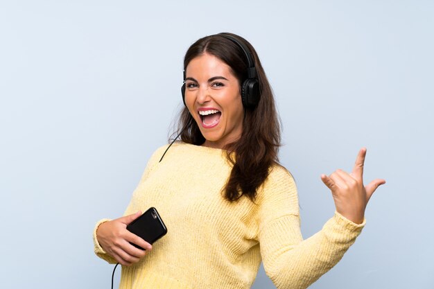 Mujer joven escuchando música con un móvil sobre pared azul aislado y bailando