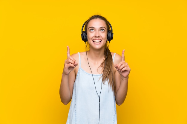 Mujer joven escuchando música sobre pared amarilla aislada apuntando hacia arriba una gran idea