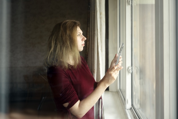 Mujer joven con un espejo en sus manos está de pie junto a la ventana