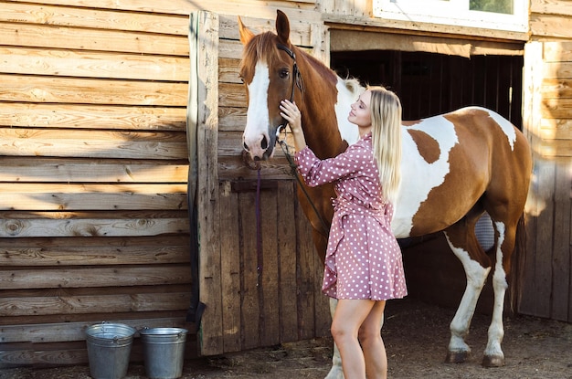Una mujer joven está acariciando y sonriendo a su caballo en un establo en un campo agrícola