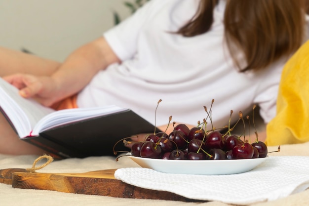 Una mujer joven está acostada con un libro en la cama cerca de un plato de cerezas.