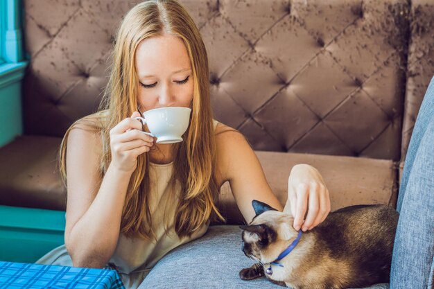 Mujer joven está bebiendo café y acariciando al gato contra el telón de fondo del sofá rayado por gatos