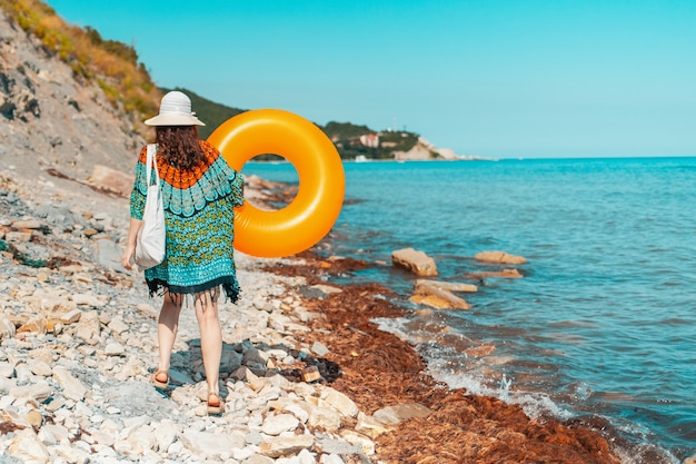 Una mujer joven está caminando sobre una playa salvaje rocosa con un círculo inflable en sus manos.