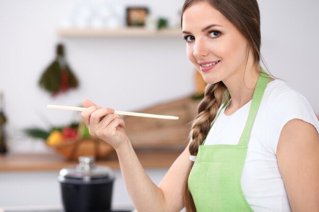 La mujer joven está cocinando en una cocina El ama de casa está probando la sopa con una cuchara de madera