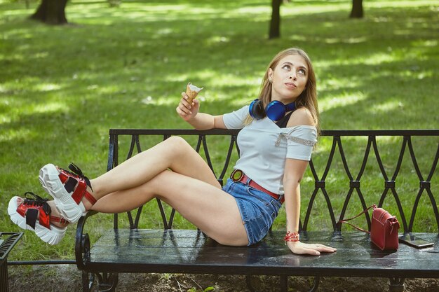 Mujer joven está comiendo helado mientras está acostado en un banco en el parque público.