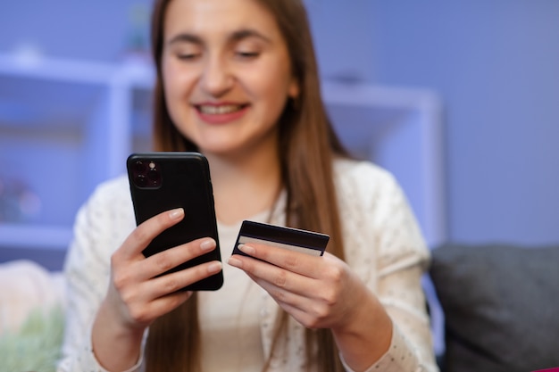 La mujer joven está comprando en línea con una tarjeta de crédito mientras está sentado en el sofá de la sala de estar. Las mujeres usan teléfonos inteligentes y realizan transacciones en línea