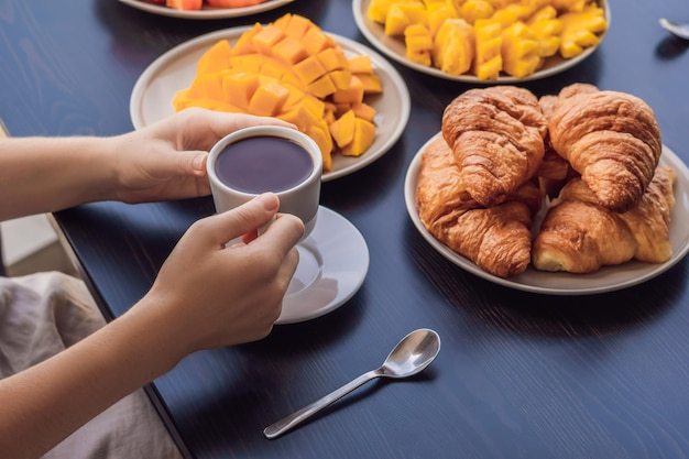 Una mujer joven está desayunando en el balcón. Mesa de desayuno con croisant de pan y fruta de café en un balcón con el telón de fondo de la gran ciudad