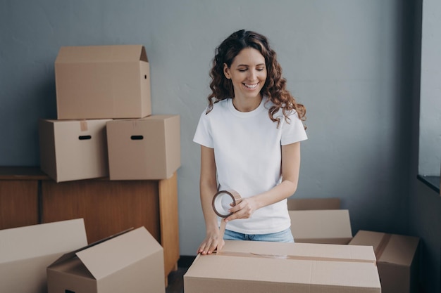 Mujer joven está empacando contenedores de cartón con cinta Trabajadora del servicio de mudanzas preparando cajas