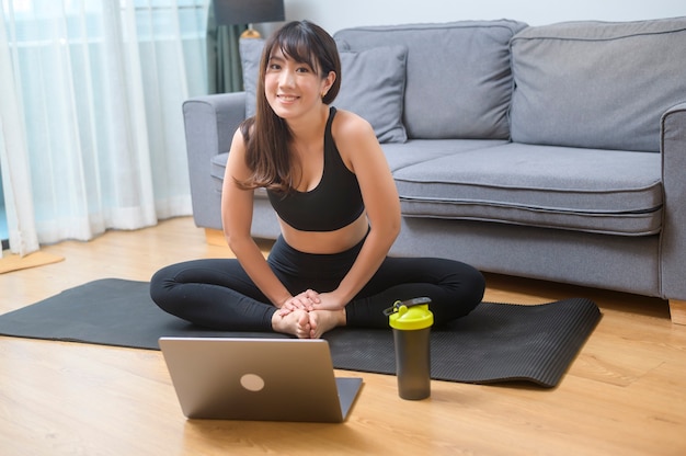 Una mujer joven está haciendo ejercicio y viendo la clase de fitness de entrenamiento en línea en la computadora portátil en la sala de estar en el hogar, el deporte, el fitness y el concepto de tecnología.