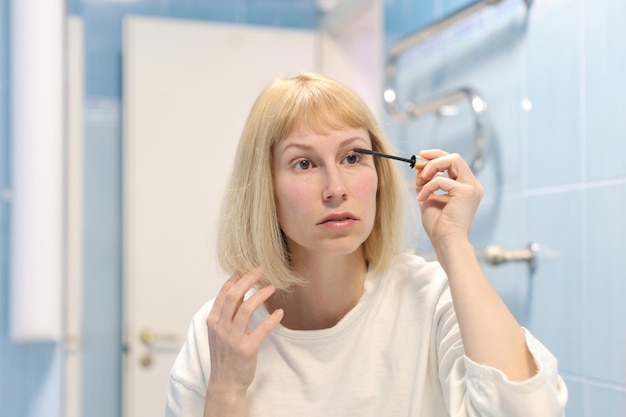 Una mujer joven está maquillando. Chica pinta sus pestañas con rímel, mirando su reflejo en el espejo de la pared. Concepto de procedimientos de maquillaje matutino femenino.