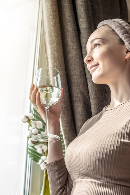 Mujer joven está de pie frente a la ventana disfrutando de una mañana soleada y sosteniendo un vaso de agua en sus manos Concepto de un comienzo agradable del día buen humor y hábitos saludables