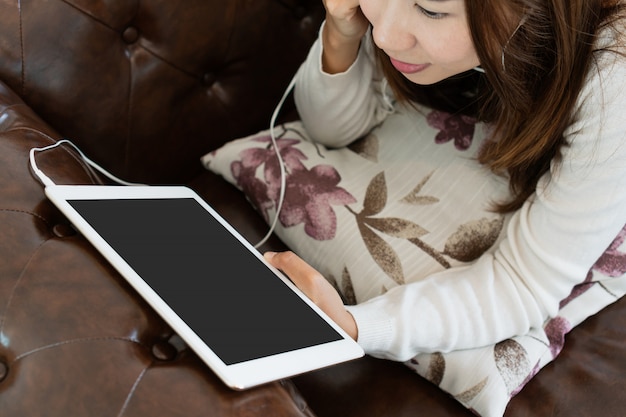 La mujer joven se está relajando en el sofá cómodo y está usando la tableta en casa.