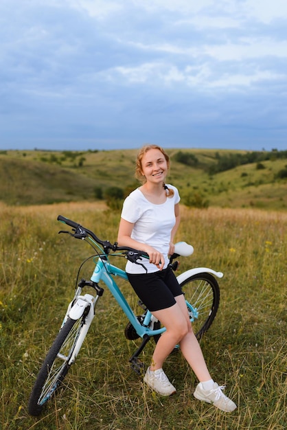 Una mujer joven está sentada en una bicicleta y mirando a la cámara en un campo contra el fondo de las colinas