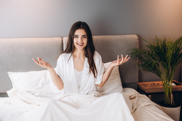 Una mujer joven está sentada en la cama con una cara de sorpresa. La modelo tiene los brazos a los lados.