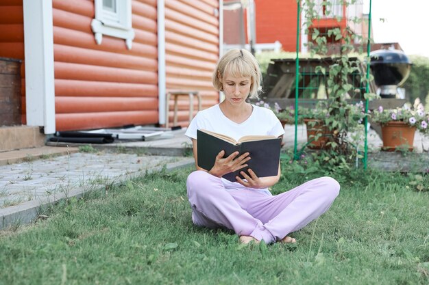 Una mujer joven está sentada en el césped del patio de su casa en un día soleado y está leyendo un libro real. Fondo de hierba y dandilion, bonito aspecto vintage.