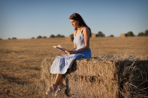 Una mujer joven está sentada en un pajar en un campo segado y leyendo un libro