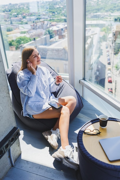 Una mujer joven está sentada en un sillón escuchando música con auriculares usando un teléfono. Una mujer bonita se relaja después de un día de trabajo relajándose en un espacio de coworking con grandes ventanas.