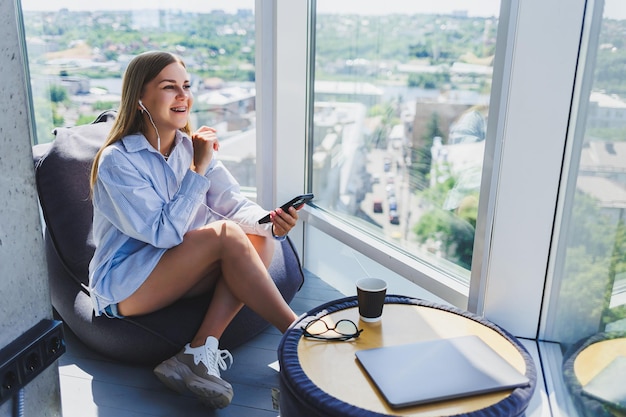 Una mujer joven está sentada en un sillón escuchando música con auriculares usando un teléfono. Una mujer bonita se relaja después de un día de trabajo relajándose en un espacio de coworking con grandes ventanas.