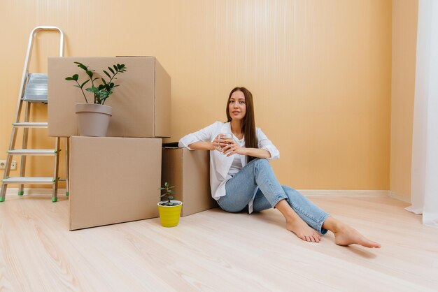 Una mujer joven está sentada en el suelo frente a las cajas y tomando café, regocijándose y disfrutando del nuevo apartamento después de mudarse. Inauguración, entrega y transporte de carga.