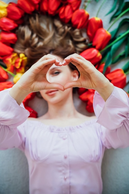 Una mujer joven está tendida en el suelo entre tulipanes rojos El concepto del 8 de marzo Día de San Valentín Retrato primaveral de una mujer Las manos de las mujeres muestran la forma de un corazón