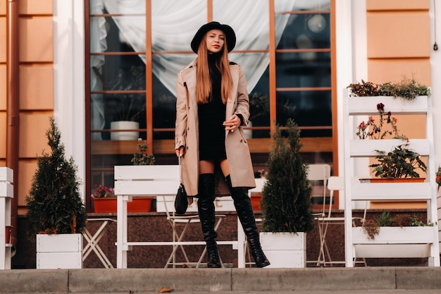 Mujer joven con estilo en un abrigo beige con un sombrero negro en una calle de la ciudad.