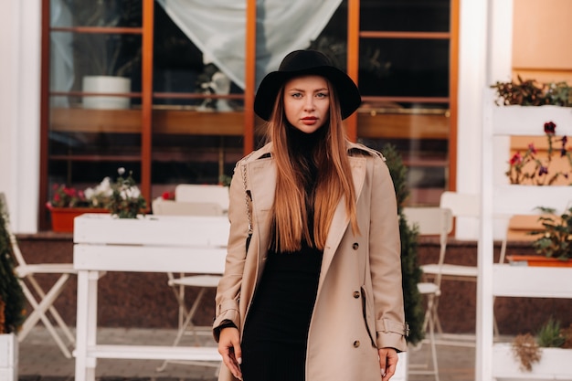 Mujer joven con estilo en un abrigo beige con un sombrero negro en una calle de la ciudad.