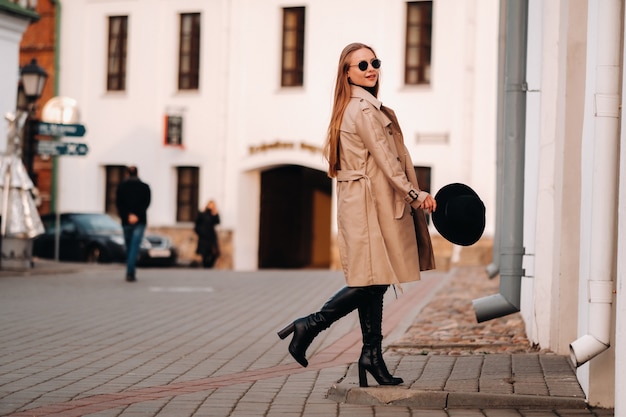 Mujer joven con estilo con un abrigo beige y un sombrero negro en sus manos y gafas en una calle de la ciudad. Moda urbana femenina. Ropa de otoño Estilo urbano.