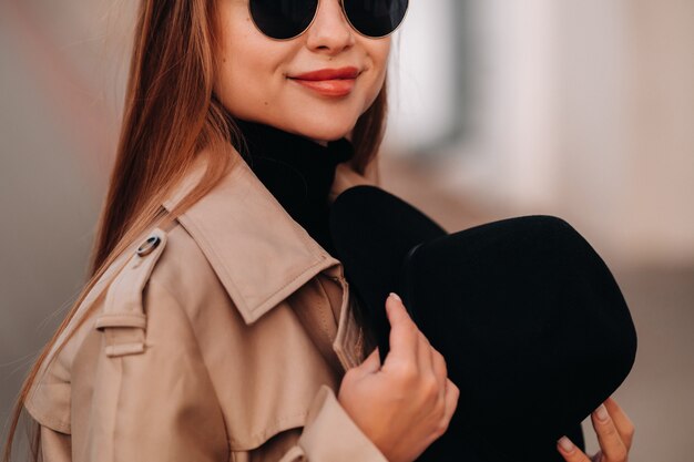 Mujer joven con estilo con un abrigo beige y un sombrero negro en sus manos y gafas en una calle de la ciudad. Moda urbana femenina. Ropa de otoño Estilo urbano.