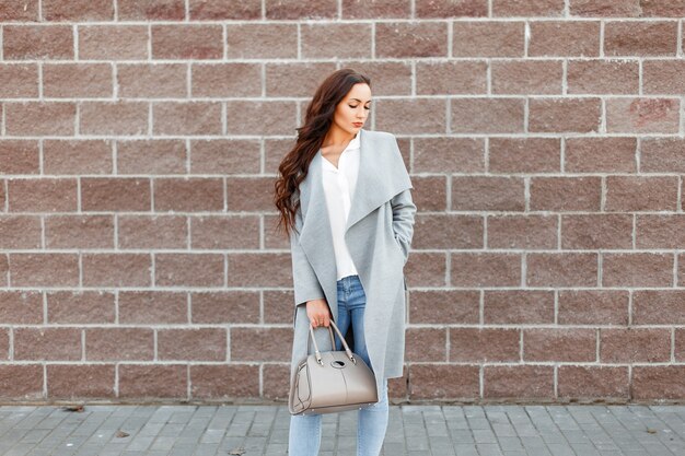 Mujer joven con estilo en un abrigo gris de moda, jeans y zapatos blancos con bolso posando cerca de una pared de ladrillos