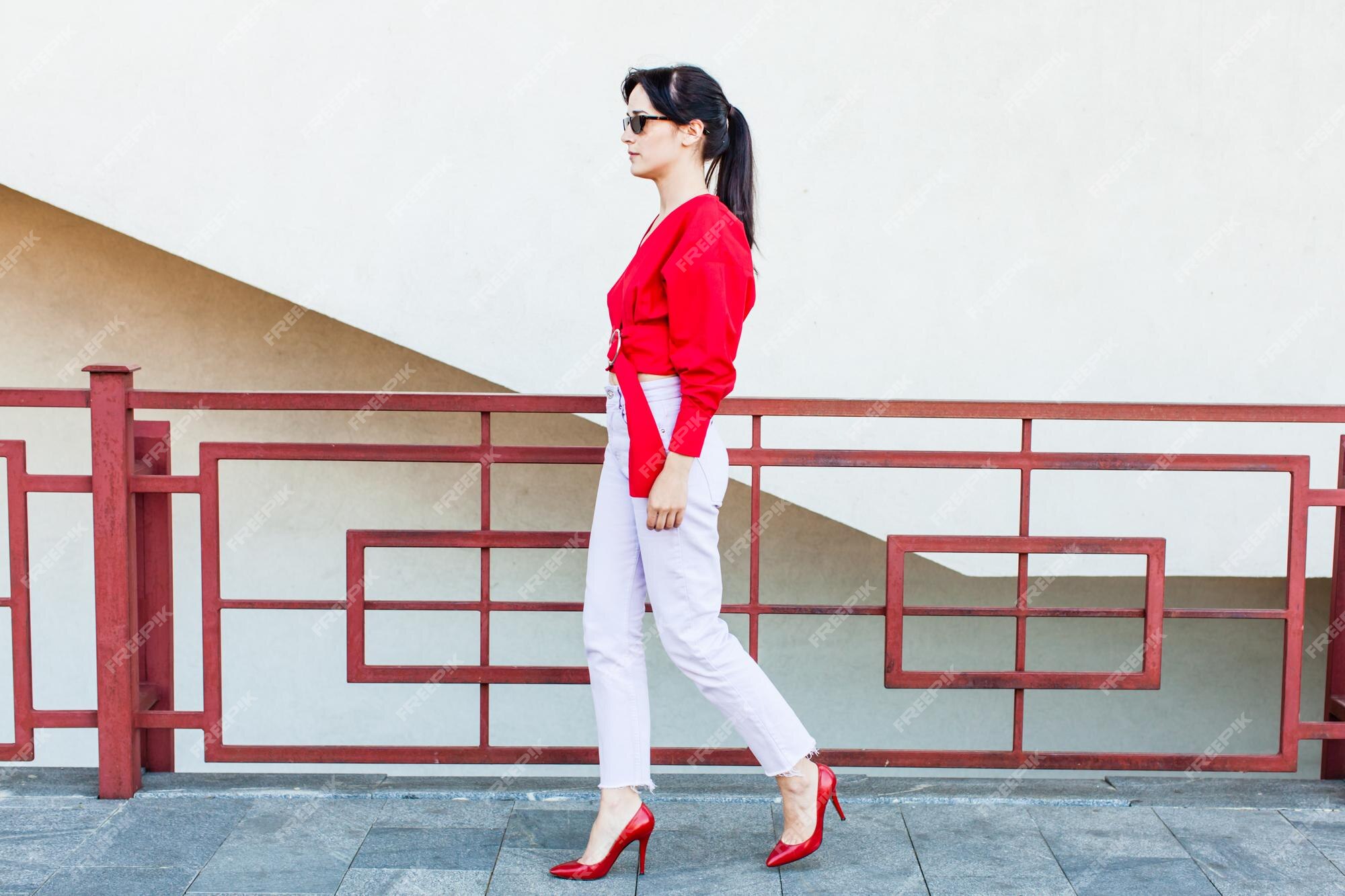 Mujer joven con estilo en una blusa roja y pantalón blanco caminando en la ciudad con zapatos de tacón alto Foto Premium