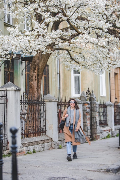 Mujer joven con estilo caminando por la calle de la ciudad en un árbol floreciente de abrigo en el estilo de vida de fondo