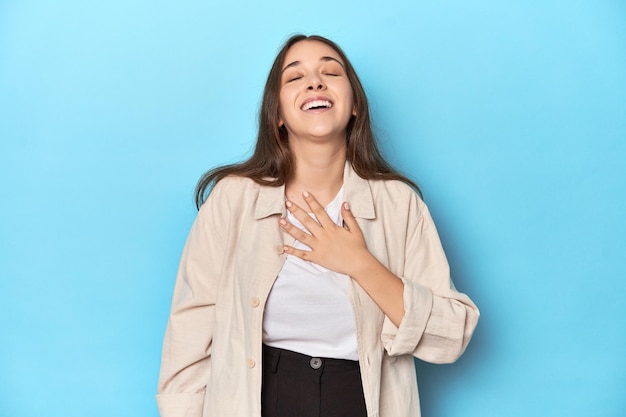 Mujer joven con estilo en una camisa sobre un fondo azul se ríe a carcajadas manteniendo la mano en el pecho