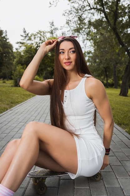 Mujer joven en estilo deportivo se sienta en una patineta y sostiene gafas de sol con la mano en un parque de la ciudad