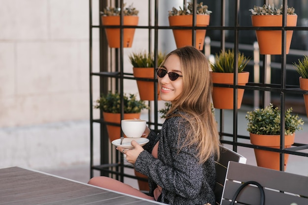 Mujer joven con estilo en gafas de sol bebe café capuchino en un café de verano mirando hacia otro lado