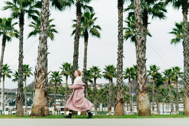 Mujer joven con estilo en un largo abrigo rosa camina en un parque junto a altas palmeras verdes cálida primavera