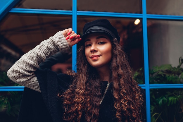 Mujer joven con estilo posando contra la ventana azul al aire libre. Traje de moda. Bella modelo con pelo rizado sonriendo