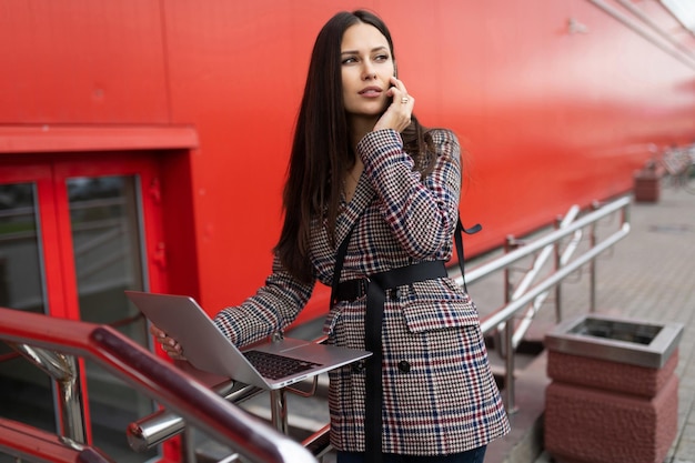 Mujer joven con estilo que trabaja en una computadora portátil en línea hablando por un teléfono móvil en el contexto de un