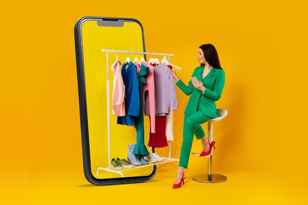 Mujer joven con estilo sentada cerca de un enorme teléfono celular y un riel de ropa usando un teléfono inteligente comprando ropa