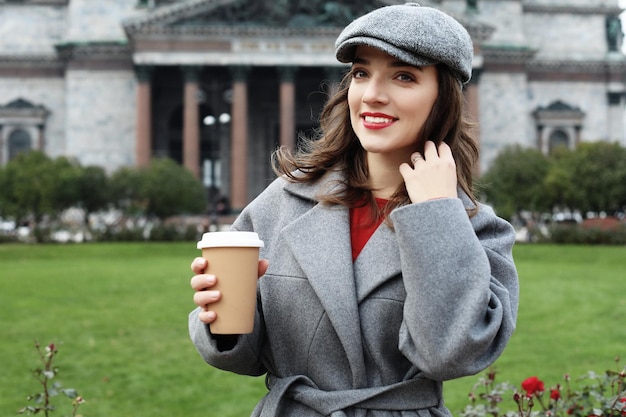 Mujer joven con estilo sonriente bebiendo café mientras camina por una calle de la ciudad
