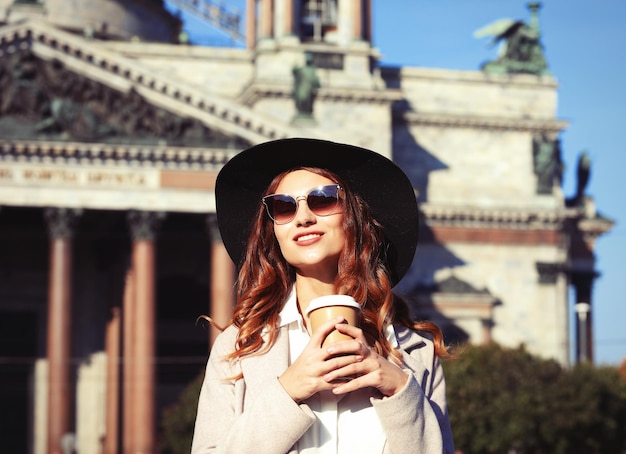 Mujer joven con estilo sonriente bebiendo café mientras camina por una calle de la ciudad