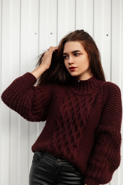 Mujer joven con estilo en un suéter burdeos posando en la ciudad cerca de una pared blanca