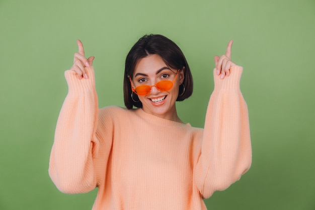 Mujer joven con estilo en suéter de melocotón casual y gafas naranjas aisladas en la pared de olivo verde emocionado señalar con los dedos índice espacio de copia