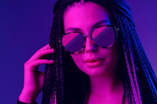 Mujer joven con estilo con trenzas con gafas de sol contra el fondo púrpura