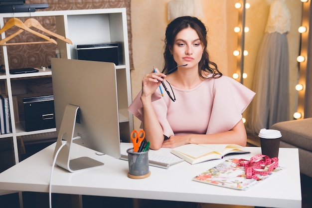 Mujer joven con estilo en vestido rosa de lujo trabajando en la oficina en la computadora