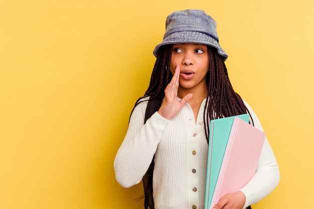 Mujer joven estudiante afroamericana aislada en la pared amarilla está diciendo una noticia secreta de frenado caliente y mirando a un lado