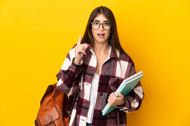 Mujer joven estudiante aislada en la pared amarilla pensando en una idea apuntando con el dedo hacia arriba