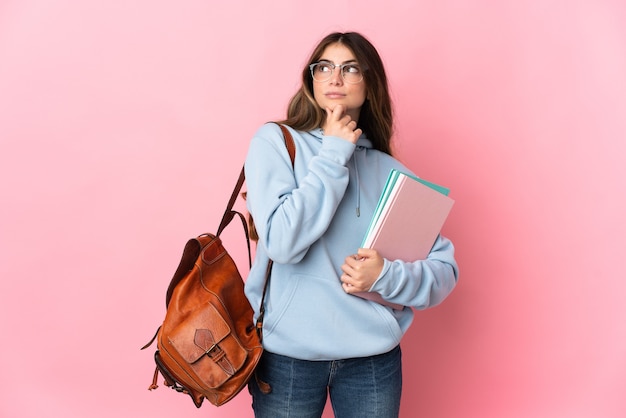 Mujer joven estudiante aislada en la pared rosa y mirando hacia arriba