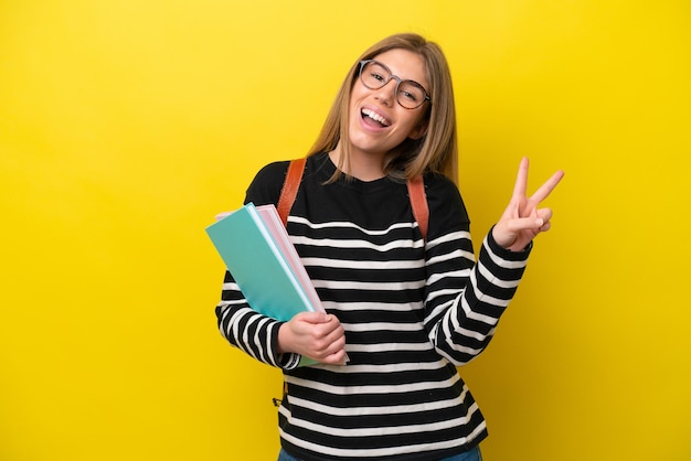 Mujer joven estudiante aislada sobre fondo amarillo sonriendo y mostrando el signo de la victoria